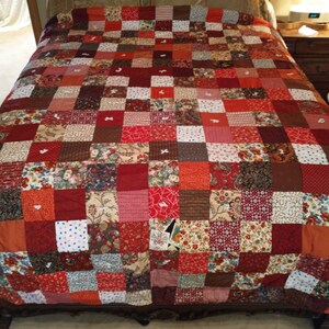 May include: A red, brown, and white patchwork quilt with a square pattern. The quilt is made of various fabrics with different patterns and textures.