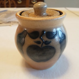 May include: A brown ceramic jar with a lid, decorated with a blue apple and leaves. The jar is approximately 4 inches tall.