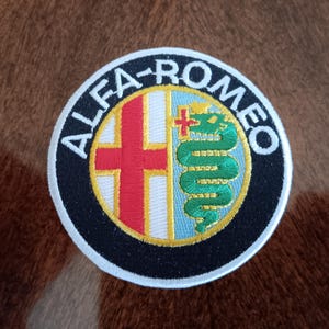 May include: Circular embroidered patch featuring the Alfa Romeo logo. The design includes a red cross on a white background, a green serpent, and the words "ALFA-ROMEO" in white lettering on a black border. The patch is likely for apparel or accessories.