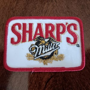 May include: A white embroidered patch with red stitching around the edges. The patch features the text "SHARP'S" in red with gold outlining. Below the text is a gold embroidered Miller logo with a black eagle.