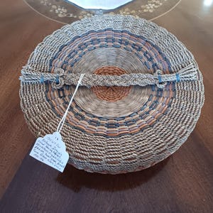 May include: Round woven basket with a lid. The basket features concentric circles of natural, orange, and dark blue hues. A woven handle is secured with blue twine. A tag hangs from the basket.