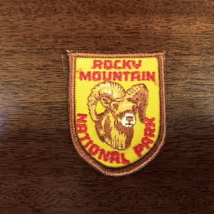 May include: A yellow and brown embroidered patch with the text "Rocky Mountain National Park" and a brown bighorn sheep illustration.