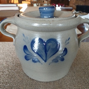 May include: A gray stoneware crock with a blue heart and floral design. The crock has a lid and two handles.