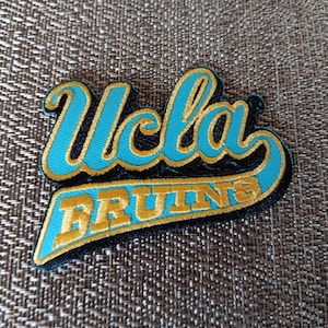 May include: Embroidered patch featuring the text "UCLA BRUINS" in a stylized script. The letters are in a vibrant teal color with gold trim, set against a black background. The patch is likely for apparel or accessories.