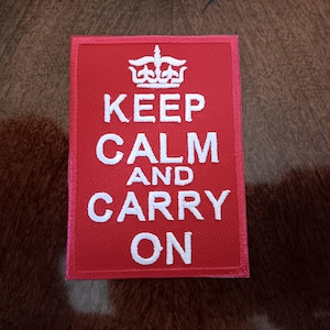 May include: A red rectangular patch with the words "KEEP CALM AND CARRY ON" in white block letters. A white crown is above the words "KEEP CALM". The patch is likely an iron-on patch or sew-on patch.