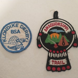 May include: Two embroidered patches. One is round with "KLONDIKE DERBY BSA" and an igloo scene in blue and white. The other is a black, stylized shape with "AMAQUONSIPPI TRAIL" and a landscape scene with trees and a campfire.