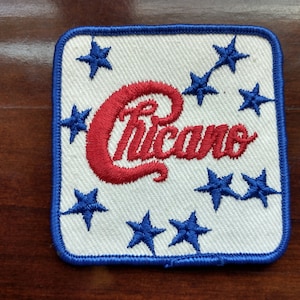 May include: Embroidered patch with the word "Chicano" in red script. The patch is square with a white background, blue border, and blue stars. The stars are arranged around the word.