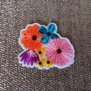 May include: Embroidered floral patch featuring an orange flower, a teal flower, a pink flower, purple leaves, and yellow flowers with black centers. The patch has a white border and is set against a textured brown background.