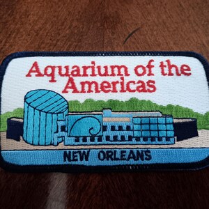 May include: A rectangular embroidered patch with a dark blue border. The white background features the red text Aquarium of the Americas above a blue building illustration, green trees, and the blue text NEW ORLEANS at the bottom edge.