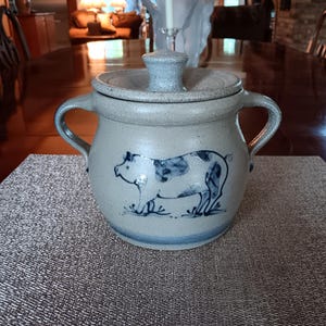 May include: A light gray stoneware crock with two side handles and a matching lid. The front of the vessel features a hand-painted blue illustration of a pig standing on grass. The base of the crock is accented with a horizontal blue band.