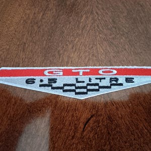 May include: A rectangular patch with the text "GTO 6.5 LITRE" in white lettering. The top border is red, the center is light gray, and the bottom features a black and white checkered pattern.