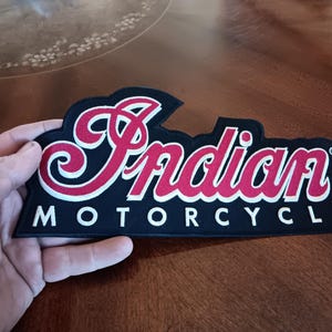 May include: Black and red embroidered patch with the text "Indian MOTORCYCLE" in white and red script. The patch has a black border and is held by a hand.