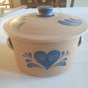 May include: Tan ceramic casserole dish with a lid, featuring blue heart and swirl designs. The dish has two handles and a blue knob on the lid. A decorative kitchenware item.