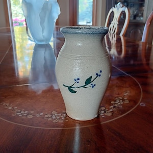 May include: A beige ceramic vase with a blue rim and a floral design. The vase has a slightly textured surface and a curved shape. The floral design features green leaves and blue berries. The vase is sitting on a wooden table.