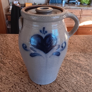 May include: A blue and white ceramic crock with a heart design and a lid. The crock is hand-painted with a blue heart and floral accents.