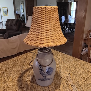 May include: A decorative lamp with a woven wicker lampshade and a ceramic base. The base is a light blue color with a blue rooster design. The lamp is sitting on a speckled countertop.