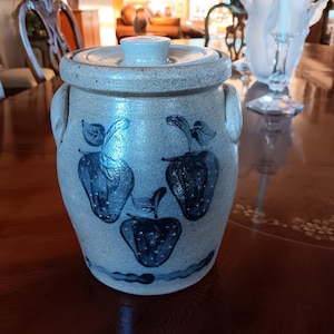 May include: A vintage stoneware crock with a lid, featuring a hand-painted design of stylized blue strawberries. The crock has two small handles and a light blue-gray color. This antique kitchenware adds a touch of rustic charm to any home.