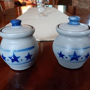 May include: Two matching ceramic jars with lids. The jars are light gray with blue accents, including a band and star designs. The lids are topped with a blue knob.