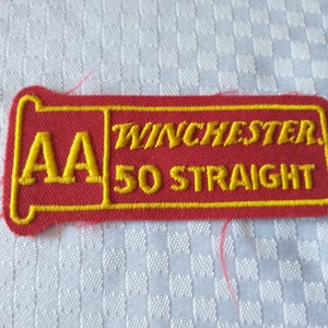 May include: A rectangular red patch with yellow embroidered text and border. The text reads "AA WINCHESTER 50 STRAIGHT". The patch has a stylized design with the letters "AA" on the left side.