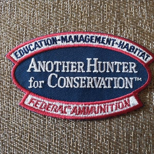 May include: A navy blue patch with white embroidered text and a red border. The patch reads "EDUCATION-MANAGEMENT-HABITAT" at the top and "FEDERAL AMMUNITION" at the bottom. The main text says "Another Hunter for Conservation".