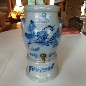 May include: A blue and white ceramic water dispenser with a bird and floral design. The dispenser has a spigot and a lid. The text on the dispenser reads "The Country Potter, Williamsburg, VA".