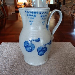 Könnte beinhalten: Ein hellgrauer Steinkrug mit einem geschwungenen Henkel und einem breiten Ausguss. Die Außenseite ist mit handgemalten kobaltblauen Blumenmotiven und einer Reihe kleiner blauer Punkte in dreieckiger Anordnung am oberen Rand verziert.