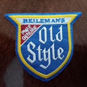 May include: A blue, yellow, and white embroidered patch with the words "Heileman's Old Style" in white lettering. The patch has a shield shape with a yellow border and a red and white section at the top.