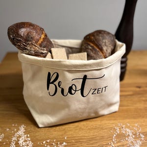 Könnte beinhalten: Ein cremefarbener Brotbeutel mit den Worten "Brot Zeit" in schwarzer Schrift. Der Beutel enthält zwei Brotlaibe und mehrere Brotstücke. Der Beutel steht auf einer Holzoberfläche.