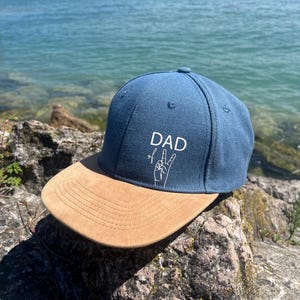 Könnte beinhalten: Eine blaue Baseballkappe mit einem flachen Schirm in beiger Wildlederoptik. Auf der Vorderseite befindet sich der weiße Schriftzug DAD über einer weißen Strichzeichnung einer Hand, die ein Victory-Zeichen zeigt. Die Kappe hat eine strukturierte Krone.