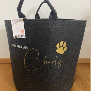 Könnte beinhalten: Eine dunkelgraue Filz-Aufbewahrungstasche mit goldener Beschriftung "HUNDEKRAM VON Charly" und einem goldenen Pfotenabdruck. Die Tasche hat zwei Henkel und ist perfekt zum Verstauen von Tierbedarf.