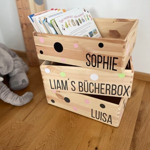 Könnte beinhalten: Gestapelte Holzkisten mit Punkten und den Namen Sophie, Liam's Bücherbox und Luisa. Bücher befinden sich in der oberen Kiste. Im Hintergrund befindet sich eine hölzerne Wachstumstabelle.