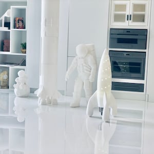Pode incluir: Uma figura de astronauta branca, uma lâmpada branca em forma de foguete e uma maquete de foguete branca estão em um chão branco. A lâmpada em forma de foguete tem uma luz dentro. A figura de astronauta está de pé com os braços ao lado do corpo. A maquete de foguete é alta e fina. O chão é brilhante e reflexivo.