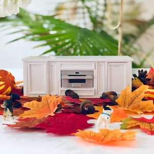 May include: A white miniature kitchen cabinet with a silver oven door. The cabinet is surrounded by fall-themed decorations, including orange and yellow gourds, brown acorns, and red and orange maple leaves.