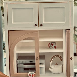 May include: White miniature kitchen cabinet with two upper cabinets, a lower cabinet with a glass door, and a side cabinet with a sliding door. The lower cabinet has two shelves and a small oven. The side cabinet has one shelf. There are two mugs and a teapot on the shelves.