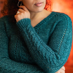 以下が含まれることがあります： ケーブル編みのパターンとVネックのターコイズブルーのニットセーターです。