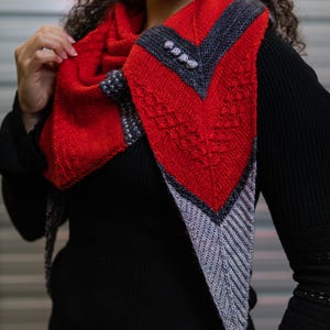 May include: A red and gray knitted shawl with a geometric design. The shawl features a vibrant red central panel with a textured pattern, bordered by gray and dark gray sections. Decorative buttons accent the design. The shawl is worn over a black top.