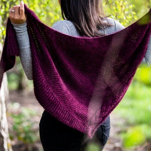 Könnte beinhalten: Eine Frau trägt einen burgunderfarbenen Strick-Schal mit lockerer, offener Maschenstruktur. Der Schal ist über ihre Schultern gelegt und sie hält ihn mit einer Hand.