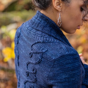 May include: A blue knitted cardigan with a unique cable knit design on the back. The cardigan has a shawl collar and long sleeves.