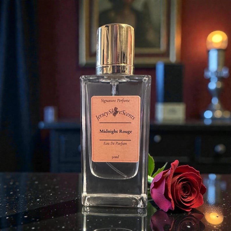 May include: A clear rectangular perfume bottle with a gold cap, labeled "Jersey Shore Scents Midnight Rouge Eau De Parfum." A dark red rose lies next to the bottle. The bottle contains 50ml of perfume.