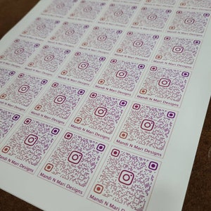 May include: A sheet of white paper with multiple stickers featuring a purple and pink Instagram QR code with the text "Mandi N Mari Designs" repeated on each sticker.