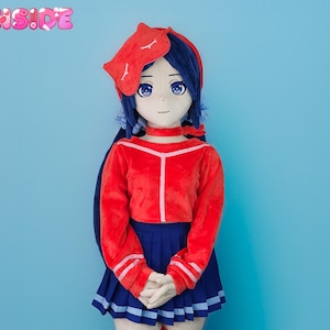 May include: A large plush doll in a sailor-inspired outfit. The doll has blue hair, a red top, a blue pleated skirt, and red and white striped thigh-high socks. The doll is wearing a red sleep mask and blue shoes. The words "PLUSH DOLL" are visible.