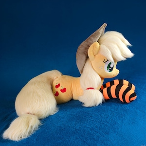 Può includere: Un peluche a forma di pony in posizione sdraiata, con corpo giallo chiaro, criniera e coda bianche e cappello marrone. Il pony ha un design a mela rossa sul fianco e un accessorio a righe arancioni e nere. Lo sfondo è blu.