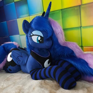 May include: A plush toy of a blue pony with a long, flowing purple and blue mane and tail. The pony has a black and white crescent moon design on its chest and is wearing blue and black striped leg warmers. The toy is lying on a light-colored surface.