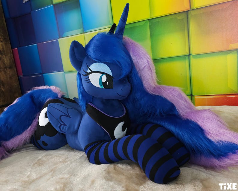 May include: A plush toy of a blue pony with a long, flowing purple and blue mane and tail. The pony has a black and white crescent moon design on its chest and is wearing blue and black striped leg warmers. The toy is lying on a light-colored surface.