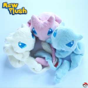 May include: Three plush toys in the shape of the Pokemon Mew. The toys are white, pink, and blue. The toys are arranged in a circle, with the white toy in the front and the pink and blue toys behind it. The toys have embroidered eyes and mouths.