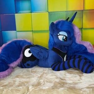 May include: A plush toy of a blue pony with a long, flowing mane and tail in shades of purple. The pony has large, expressive eyes and is lying on a beige surface. The background features a colorful, geometric wall.