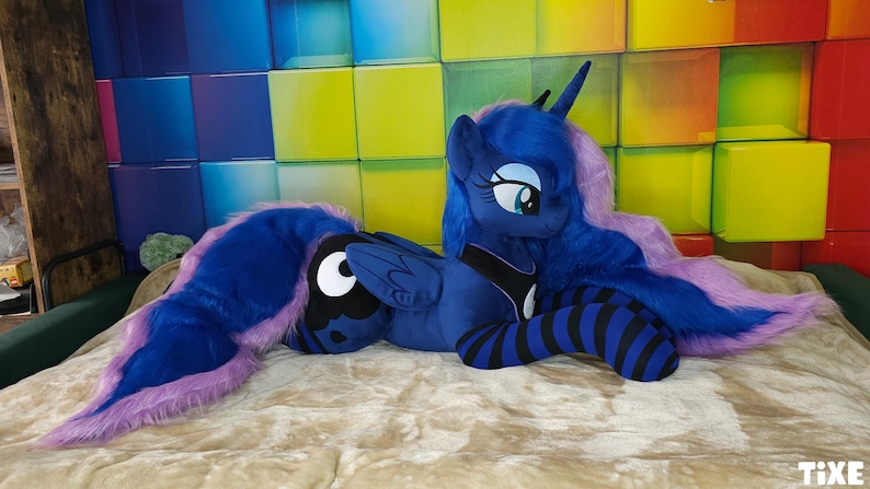 May include: A plush toy of a blue pony with a long, flowing mane and tail in shades of purple. The pony has large, expressive eyes and is lying on a beige surface. The background features a colorful, geometric wall.