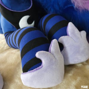 May include: Close-up of plush toy legs and hands. The legs are covered in blue and black striped fabric. The hands are light purple with a soft, fuzzy texture. The background is a light tan color.