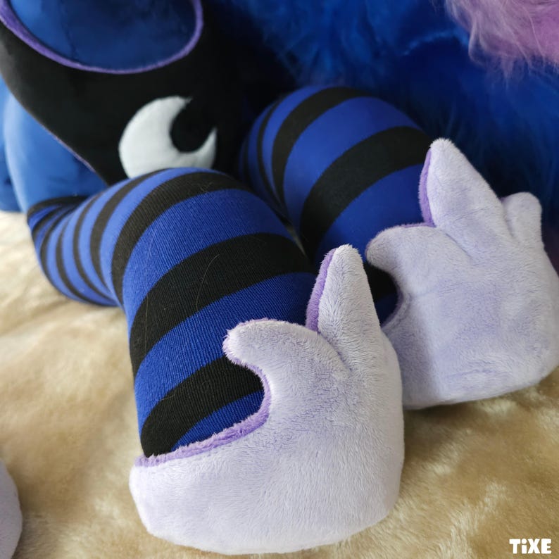 May include: Close-up of plush toy legs and hands. The legs are covered in blue and black striped fabric. The hands are light purple with a soft, fuzzy texture. The background is a light tan color.