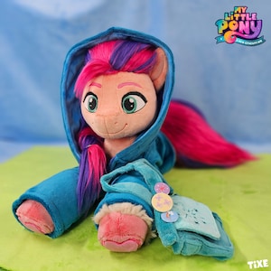 Könnte beinhalten: Ein Plüschtier eines rosa und violetten Ponys, das eine blaue Kapuzenjacke trägt. Das Pony hat eine rosa Nase und grüne Augen. Die Jacke hat ein blau-weiß gestreiftes Muster und einen weißen Pelzbesatz. Das Pony sitzt auf einer grünen Oberfläche. Der Text "My Little Pony: A New Generation" ist im Hintergrund sichtbar.